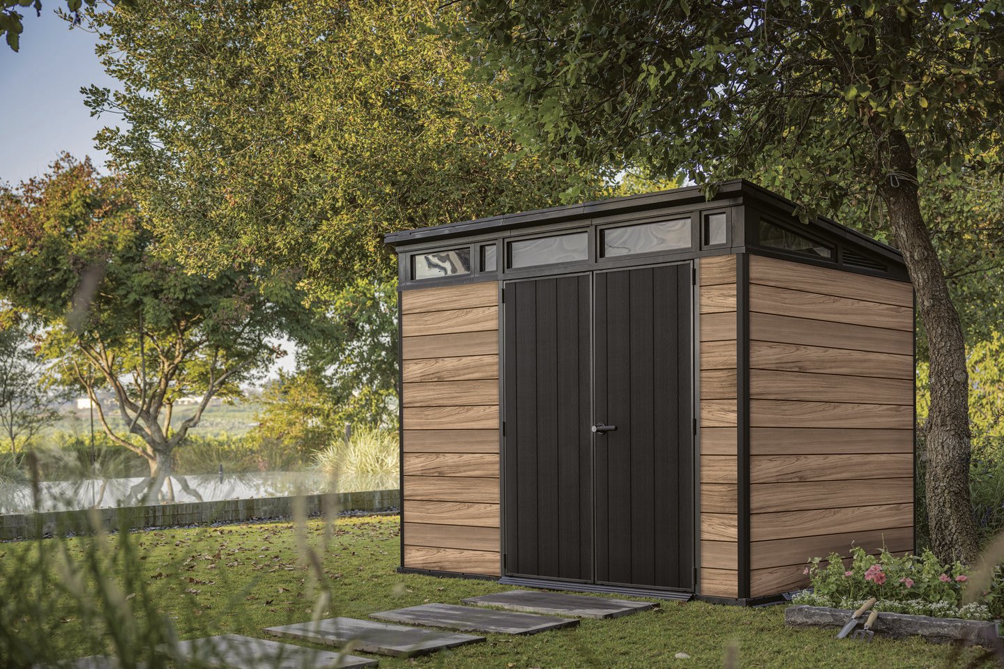 Keter tuinhuis met houtlook panelen en zwarte deur, geplaatst in een groene tuin naast een pad van stenen tegels.