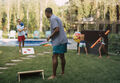 two men play cornhole while two children grab pool toys from a Keter deck box
