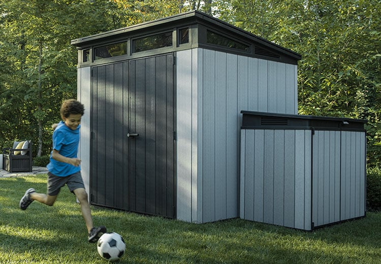 Boy kicks soccer ball past two Keter sheds